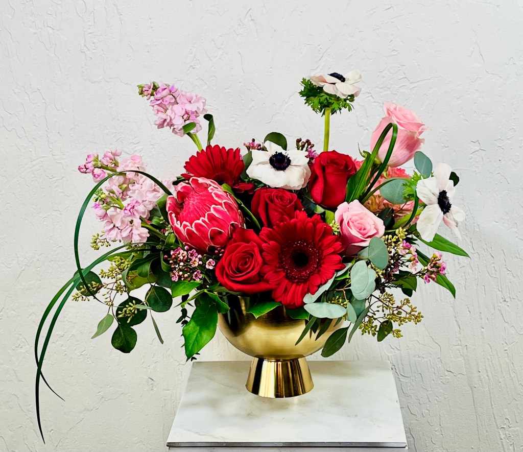 red gerbera daisies, white anemone, pink stock, pink roses, red roses, pink mink protea arranged in a gold compote designed by la paloma blanca floral designs in scottsdale, az