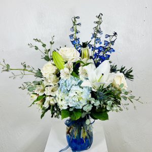 Clear vase with blue crystals in the bottom, filled with blue hydrangea, blue delphinium, white roses, blue thistle, and white oriental lilies designed by La Paloma Blanca Floral Designs