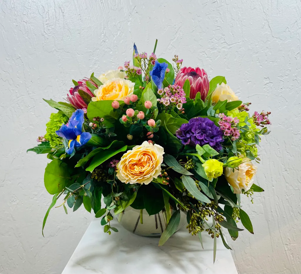 clear bubble bowl filled with yellow garden roses, purple lisianthus, pink mink protea, blue iris, green mini hydrangea, and pink hypericum berries designed by La Paloma Blanca Floral Designs in Scottsdale, AZ