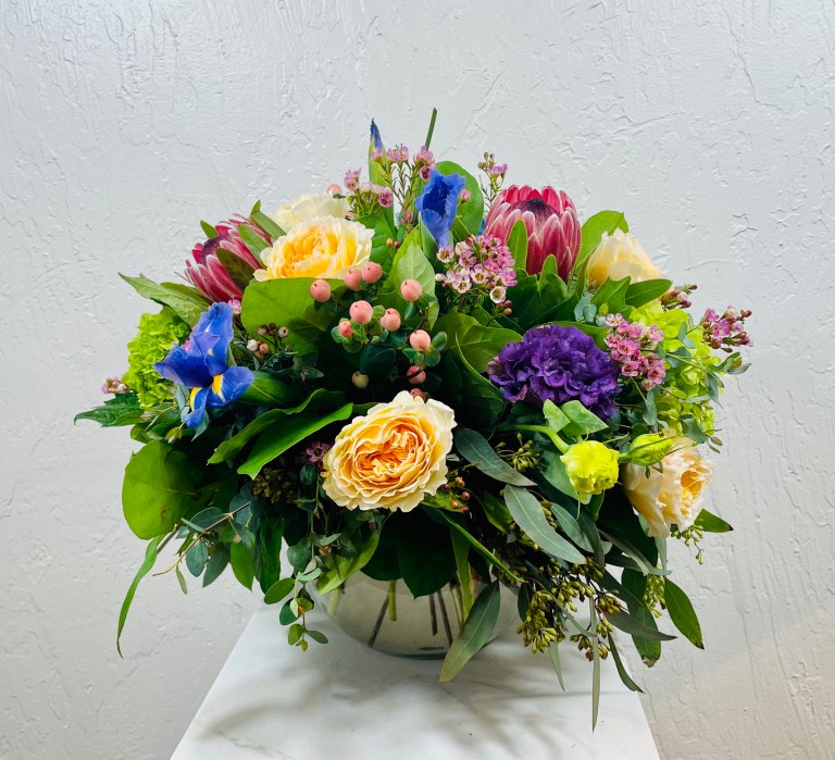 clear bubble bowl filled with yellow garden roses, purple lisianthus, pink mink protea, blue iris, green mini hydrangea, and pink hypericum berries designed by La Paloma Blanca Floral Designs in Scottsdale, AZ