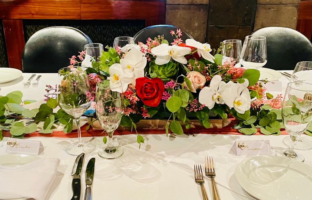 burnt orange roses, white phaelanopsis orchids, pink mink protea, champagne roses, and greens low in a glass vase with a wood bottom. on a table with white linens and silverware.