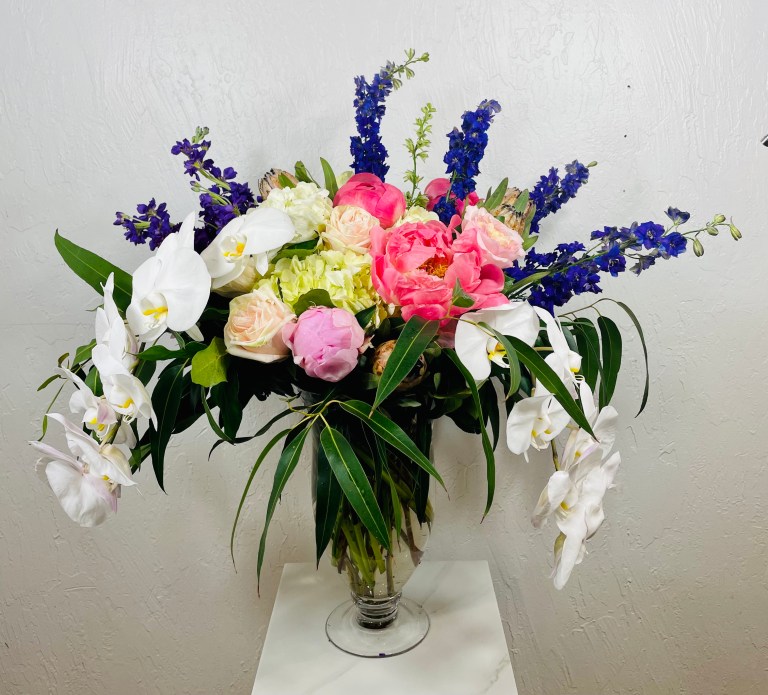 Purple larkspur, pink roses, pink peonies, white moth orchids, pink mink protea, willow eucalyptus in a pedestal vase designed by La Paloma Blanca Floral Designs in Scottsdale, AZ