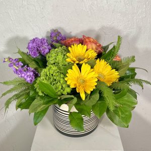 Black and white striped pot filled with green hydrangea, yellow gerbera daisies, orange roses, and lavender stock