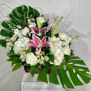 White roses, white hydrangea, white stock, white alstroemeria, and pink stargazer lilies surrounded by monstera leaves in a basket